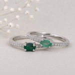 Two silver emerald rings with natural oval green emerald-cut gemstones and pavé-set clear stones, displayed on a light neutral surface with delicate white flowers in the background.