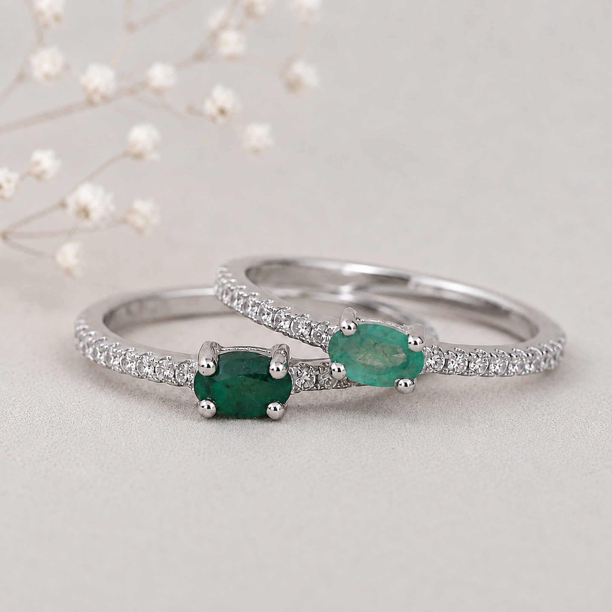 Two silver emerald rings with natural oval green emerald-cut gemstones and pavé-set clear stones, displayed on a light neutral surface with delicate white flowers in the background.