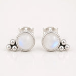 A pair of silver stud earrings featuring a round white moonstone with a subtle blue iridescent sheen, accented by three small polished silver beads on each earring, set against a clean white background.