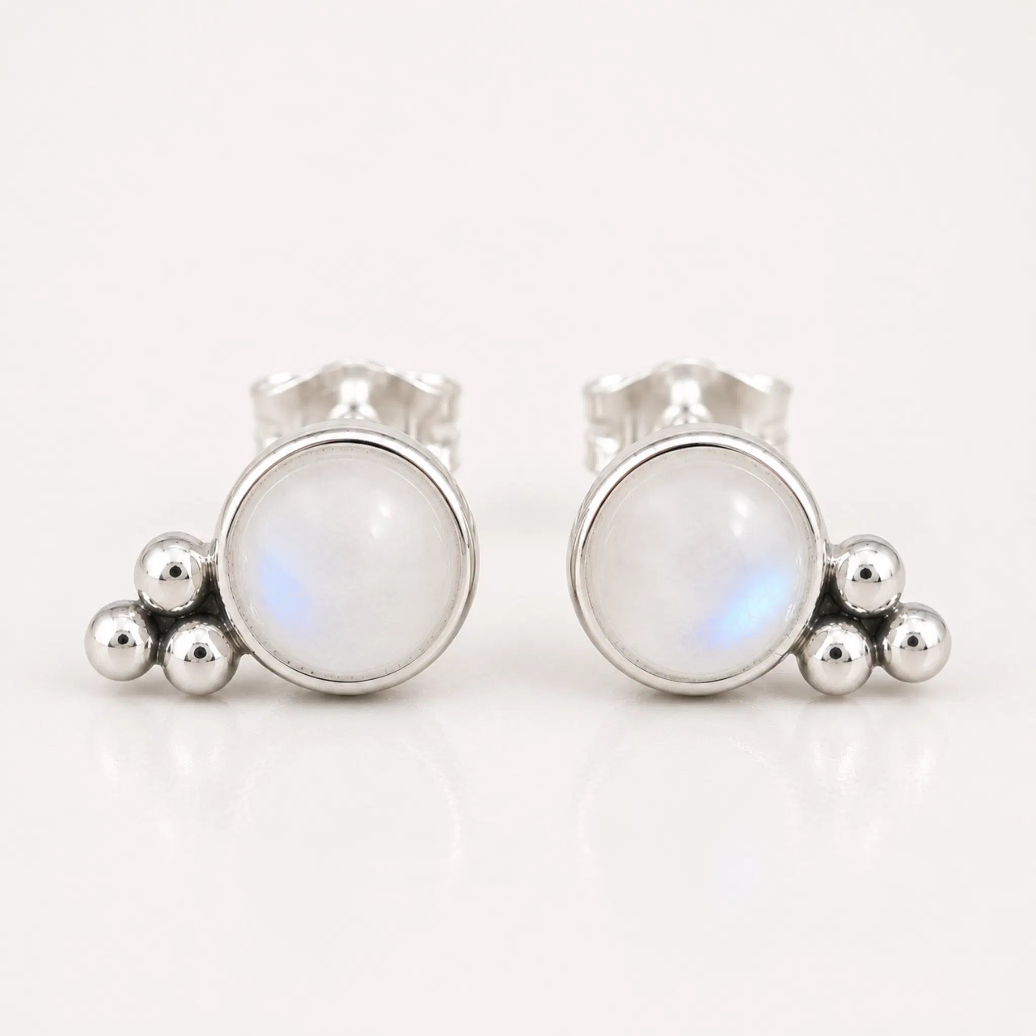 A pair of silver stud earrings featuring a round white moonstone with a subtle blue iridescent sheen, accented by three small polished silver beads on each earring, set against a clean white background.