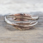 A silver wrap ring designed as two overlapping feathers, resting on a rustic wooden surface.
