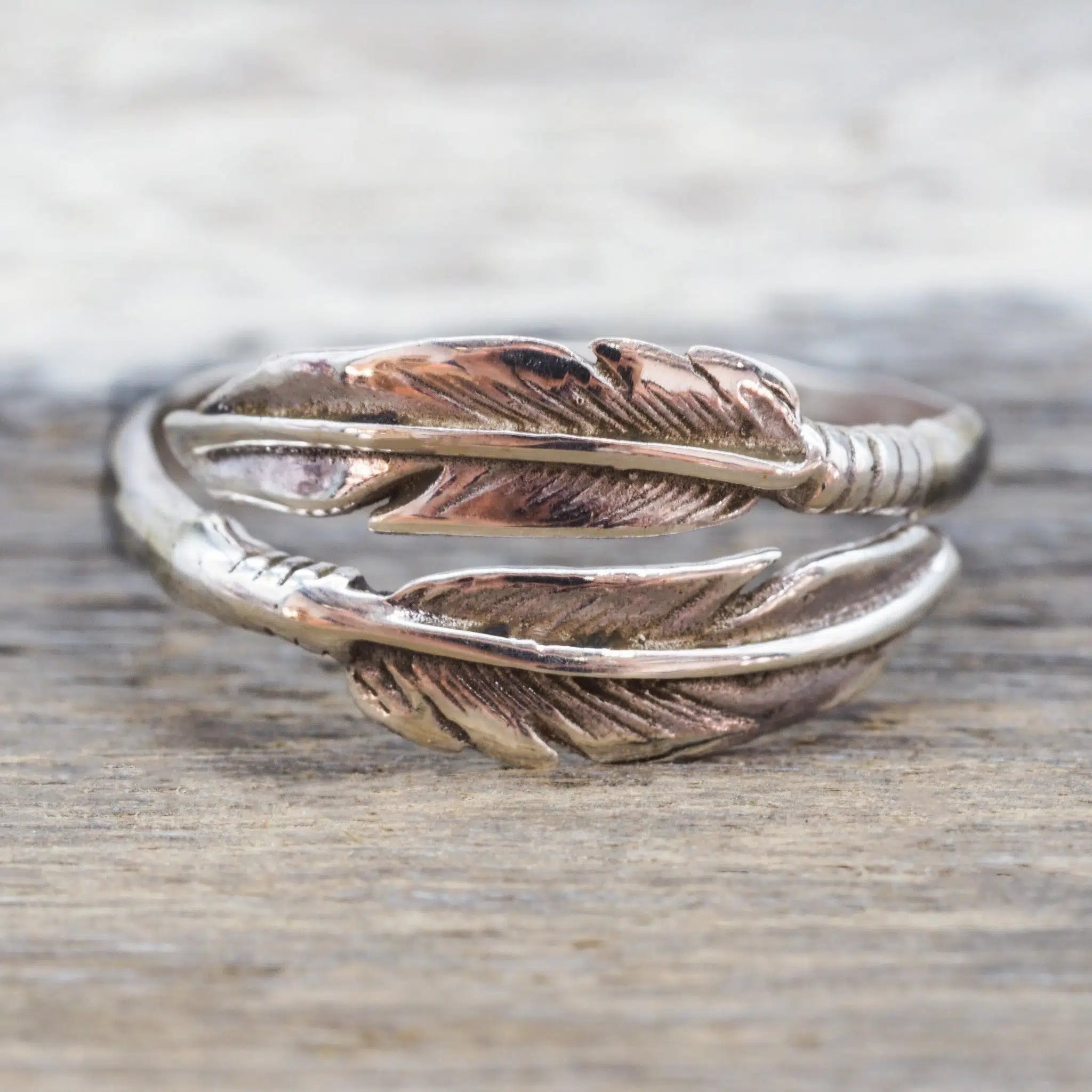 A silver wrap ring designed as two overlapping feathers, resting on a rustic wooden surface.