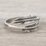 A sterling silver feather ring with detailed feather engravings, resting on a light gray wood-textured surface.