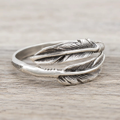 A sterling silver feather ring with detailed feather engravings, resting on a light gray wood-textured surface.
