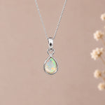 A silver october birthstone necklace featuring a teardrop pendant with a iridescent natural opal gemstone, suspended from a delicate silver chain against a soft beige background with blurred dried flowers on the right.