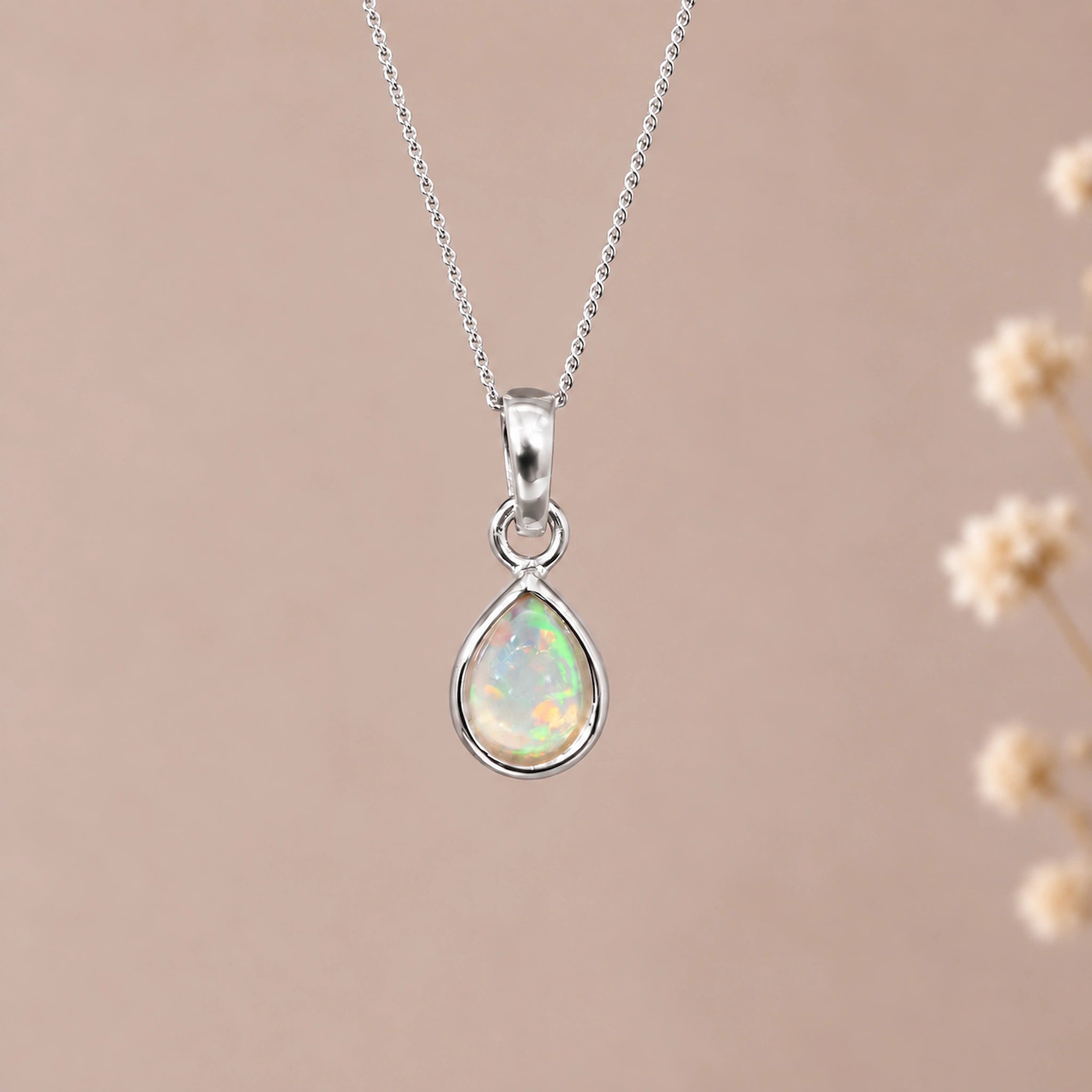 A silver october birthstone necklace featuring a teardrop pendant with a iridescent natural opal gemstone, suspended from a delicate silver chain against a soft beige background with blurred dried flowers on the right.