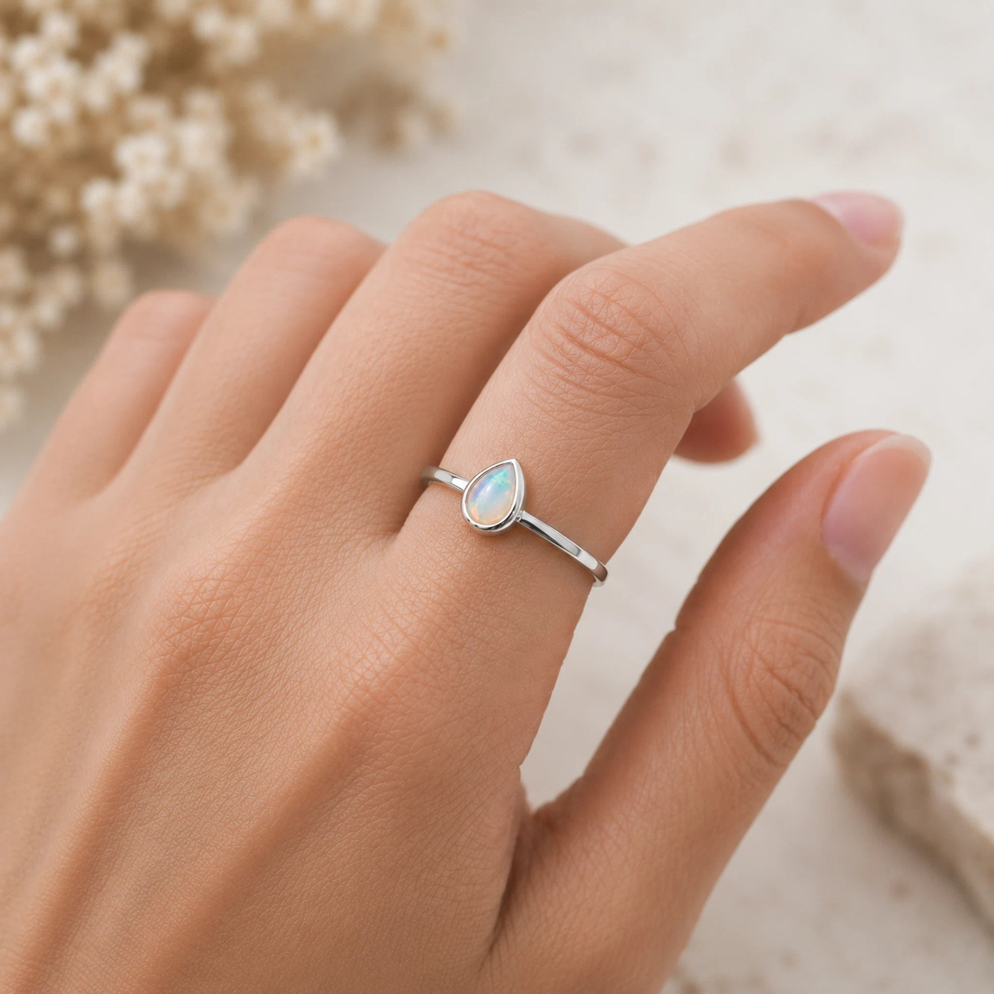 A silver ring with a teardrop-shaped iridescent opal stone, worn on a person's finger against a soft, light beige background with blurred dried flowers.
