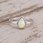 A silver teardrop ring featuring a vibrant iridescent opal gemstone, resting on a rustic, light-colored wooden surface.