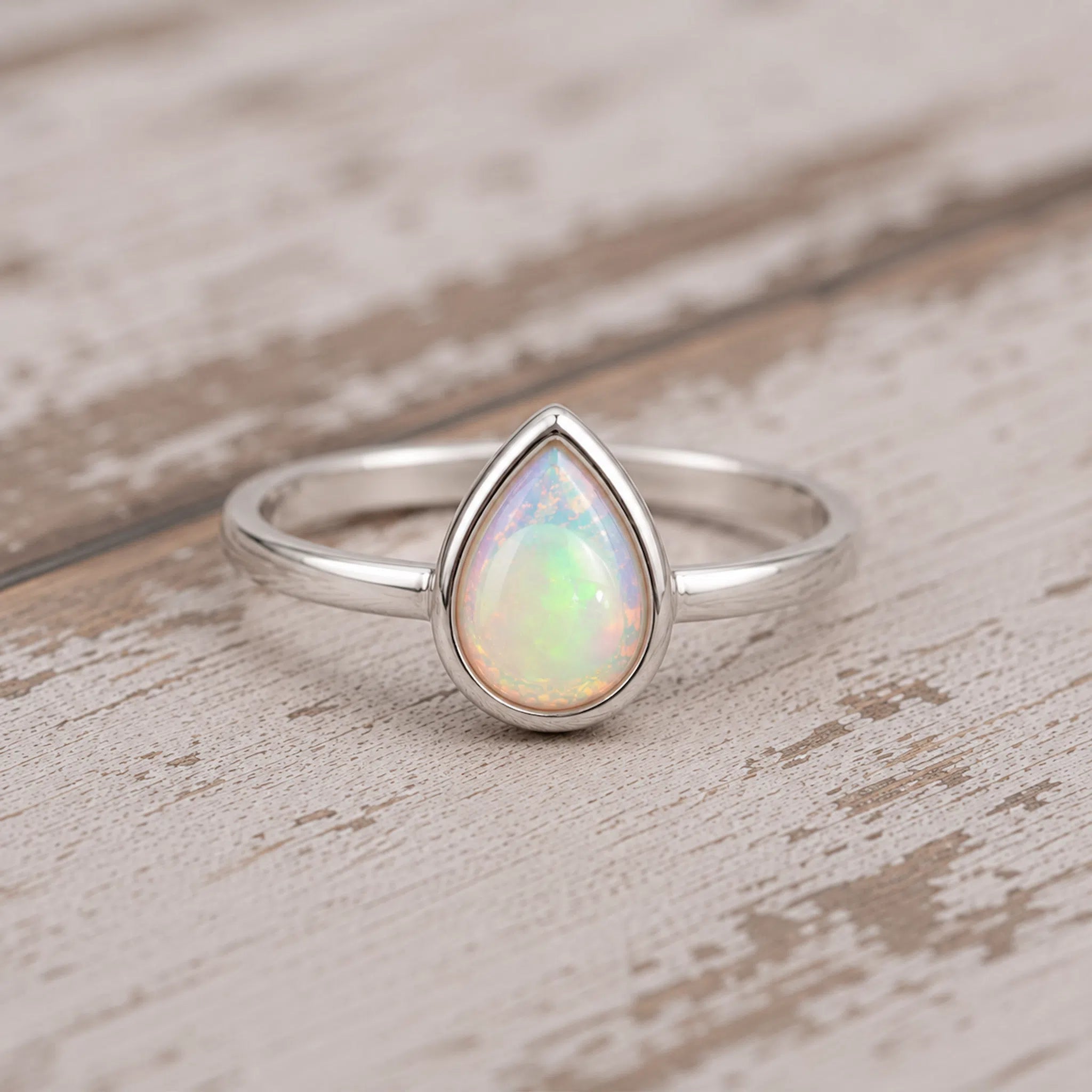 A silver teardrop ring featuring a vibrant iridescent opal gemstone, resting on a rustic, light-colored wooden surface.