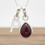Silver necklace with a teardrop-shaped red ruby gemstone pendant and letter 'P' charm on a wooden surface.