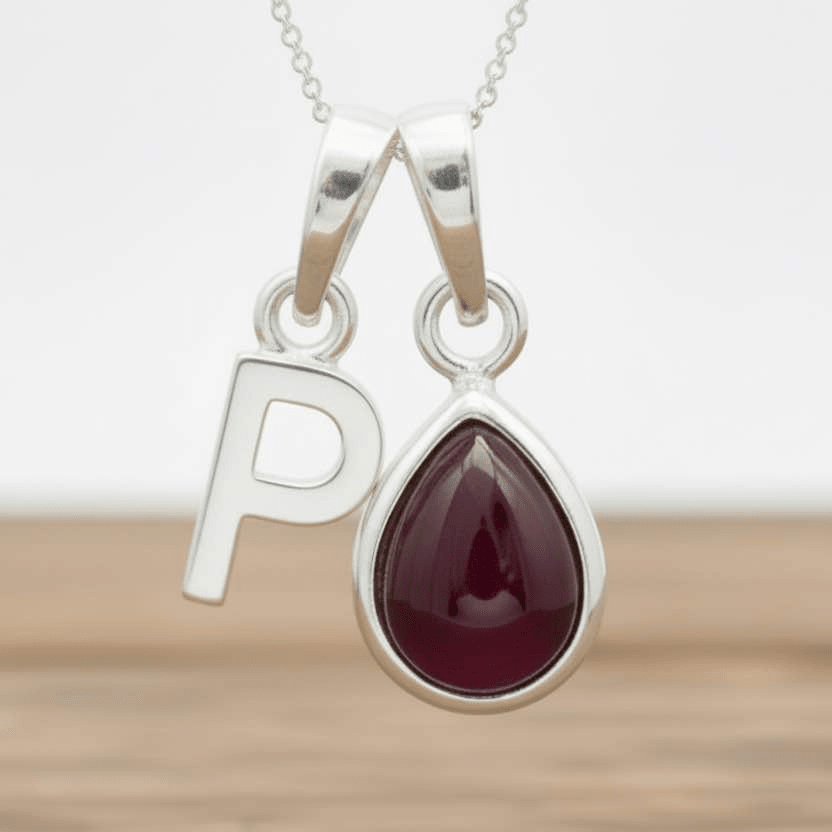 Silver necklace with a teardrop-shaped red ruby gemstone pendant and letter 'P' charm on a wooden surface.