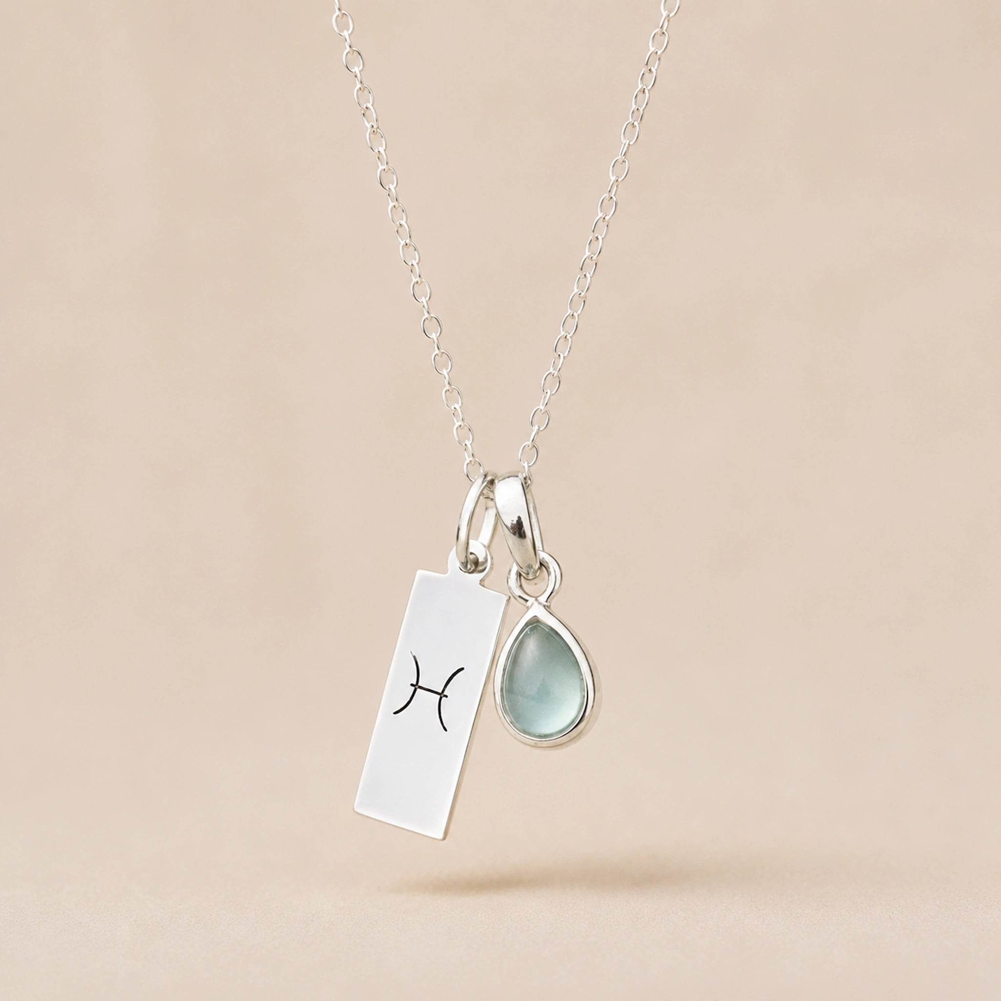 Sterling Silver Necklace with teardrop March Birthstone Natural Aquamarine pendant and a rectangle pendant wth Pisces line glyph hanging on a light background.