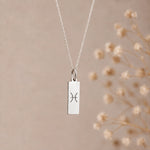 A silver pisces necklace featuring a rectangular pendant with an engraved Pisces zodiac symbol, hanging from a delicate silver chain against a soft beige background with blurred dried flowers on the right.