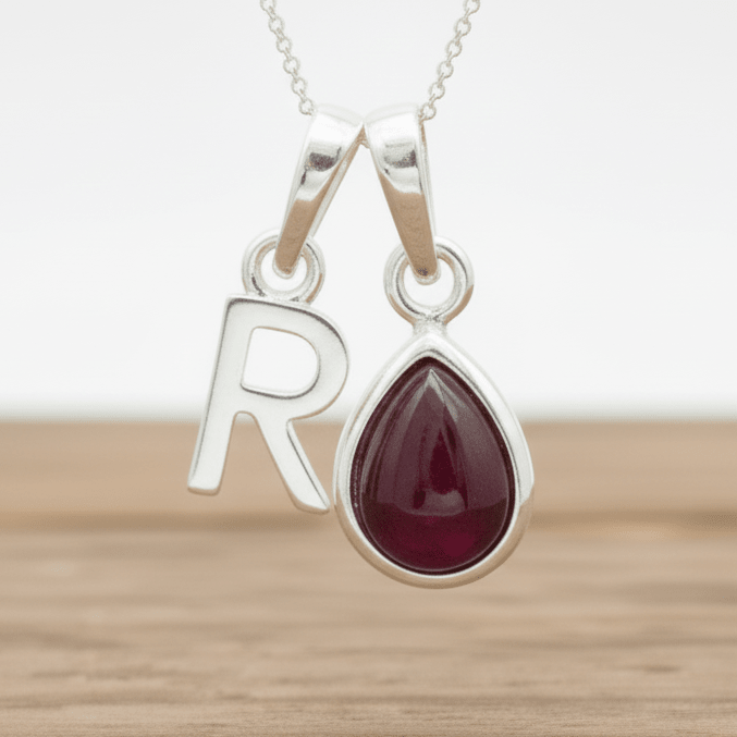 Silver necklace with a teardrop-shaped red ruby gemstone pendant and 'R' initial charm on a wooden surface.