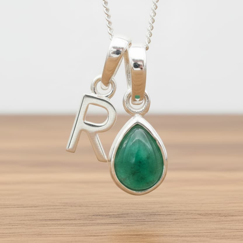 Silver Initial R necklace with a teardrop-shaped Emerald gemstone pendant hanging above a rustic wooden surface