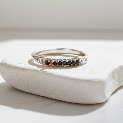 Silver ring with sapphire stones on a white stone surface next to a vase with dried plants.