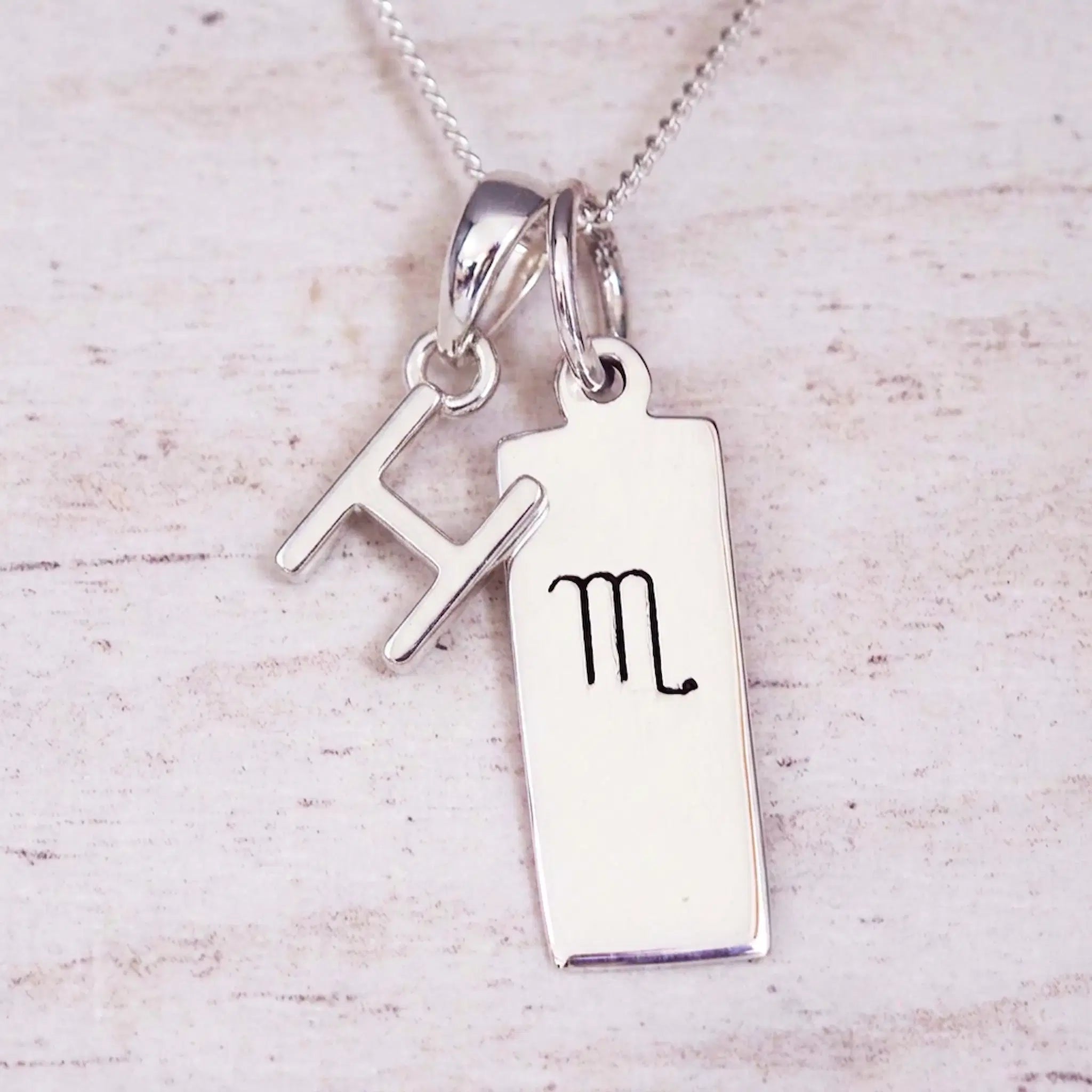 Silver Initial and Scorpio Zodiac Necklace with a letter H pendant and engraved Scorpio bar, flat lay on light wood