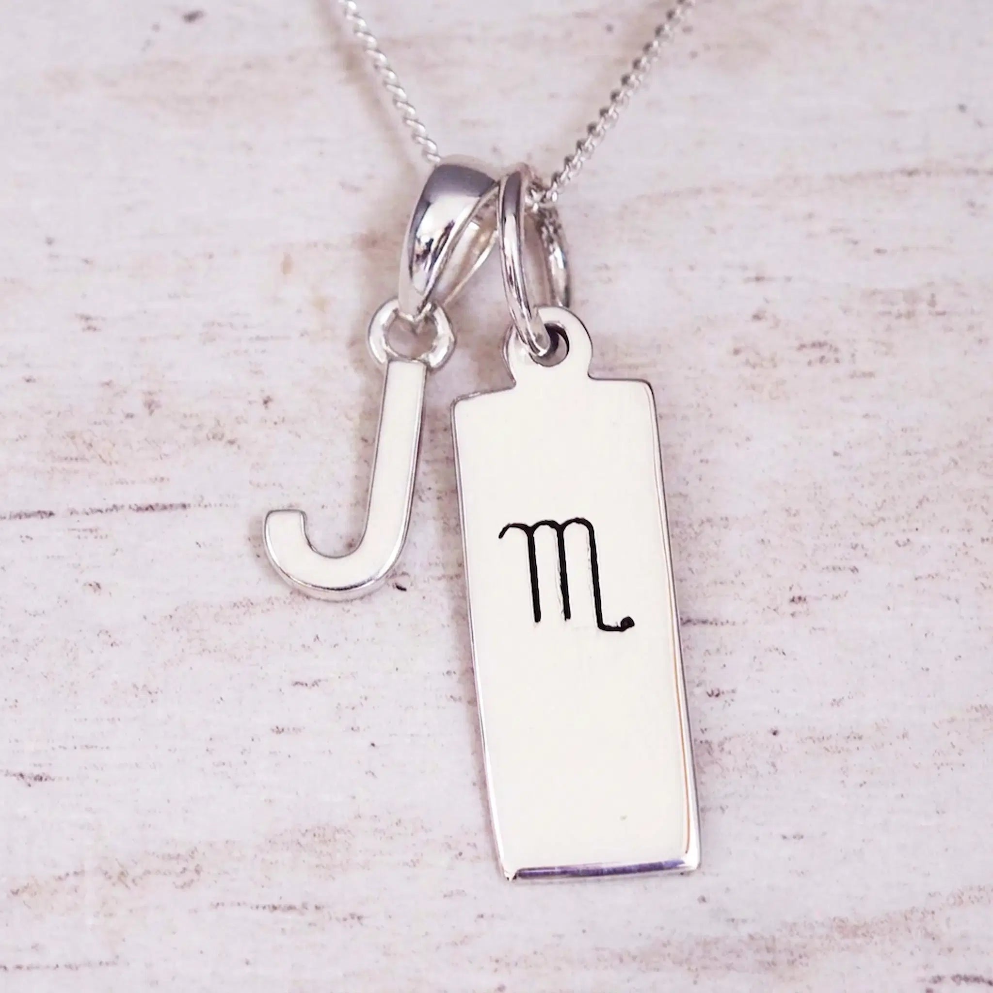 Silver Initial and Scorpio Zodiac Necklace with a letter J pendant and engraved Scorpio bar, flat lay on light wood