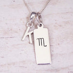 Silver Initial and Scorpio Zodiac Necklace with a letter K pendant and engraved Scorpio bar, flat lay on light wood