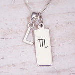 Silver Initial and Scorpio Zodiac Necklace with a letter L pendant and engraved Scorpio bar, flat lay on light wood