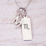 Silver Initial and Scorpio Zodiac Necklace with a letter M pendant and engraved Scorpio bar, flat lay on light wood