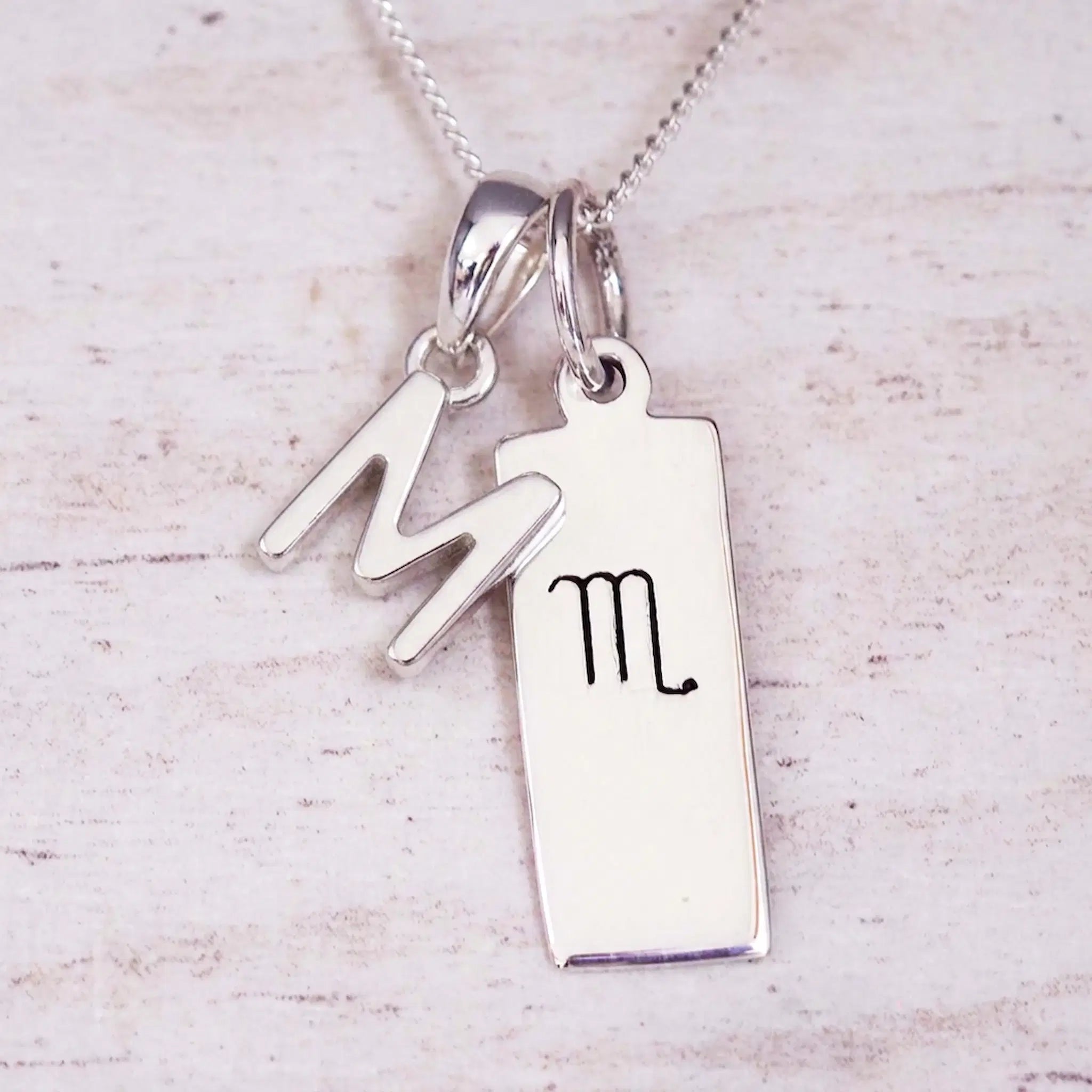 Silver Initial and Scorpio Zodiac Necklace with a letter M pendant and engraved Scorpio bar, flat lay on light wood