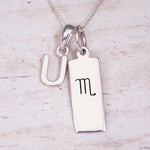 Silver Initial and Scorpio Zodiac Necklace with a letter U pendant and engraved Scorpio bar, flat lay on light wood