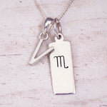 Silver Initial and Scorpio Zodiac Necklace with a letter V pendant and engraved Scorpio bar, flat lay on light wood