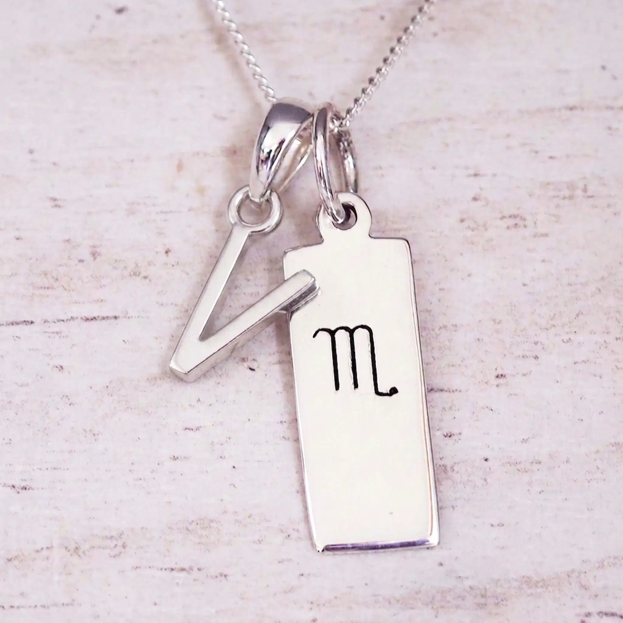 Silver Initial and Scorpio Zodiac Necklace with a letter V pendant and engraved Scorpio bar, flat lay on light wood