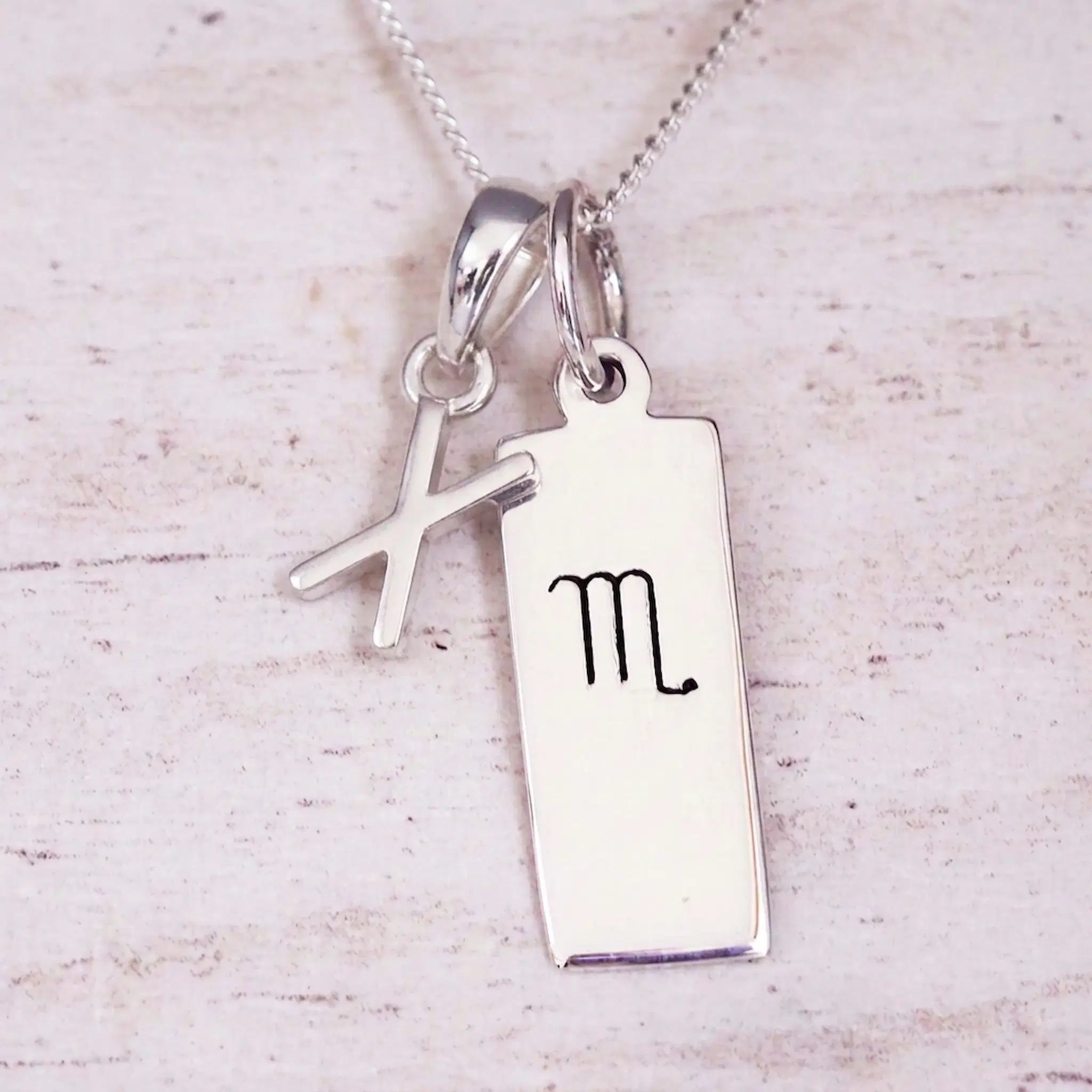 Silver Initial and Scorpio Zodiac Necklace with a letter X pendant and engraved Scorpio bar, flat lay on light wood
