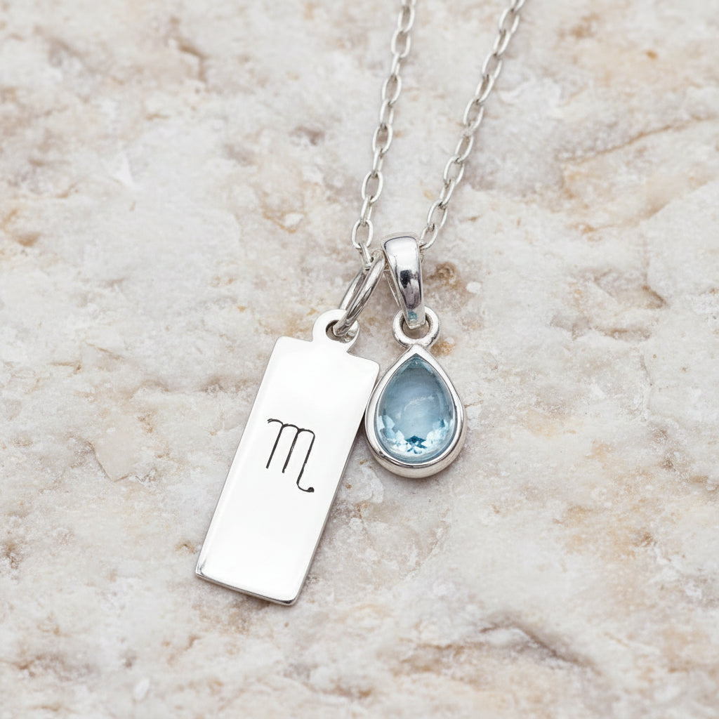 Silver necklace with Scorpio symbol pendant and a natural tear drop blue topaz gemstone pendant on a textured dark background