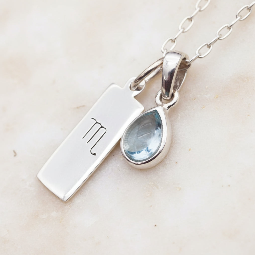 Silver necklace with Scorpio zodiac charm and teardrop-shaped natural blue topaz gemstone pendant on a textured surface.