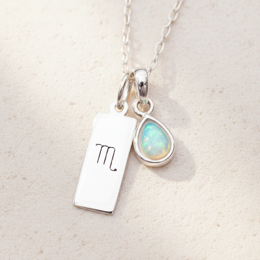 Silver necklace with Scorpio zodiac charm and teardrop-shaped natural opal pendant on a warm stone background