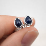 A pair of September birthstone natural sapphire earrings - silver teardrop-shaped cufflinks with dark blue cabochon stones, shown being worn on a person's fingers against a light gray background.