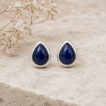 A pair of September Birthstone Natural Sapphire Earrings - silver stud earrings with teardrop-shaped dark blue lapis lazuli stones, displayed on a light beige textured stone surface with dried baby's breath flowers in the background.