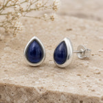A pair of September Birthstone natural Sapphire Earrings - silver teardrop stud earrings with deep blue gemstone centers, displayed on a textured beige stone surface with small white flowers in the background.
