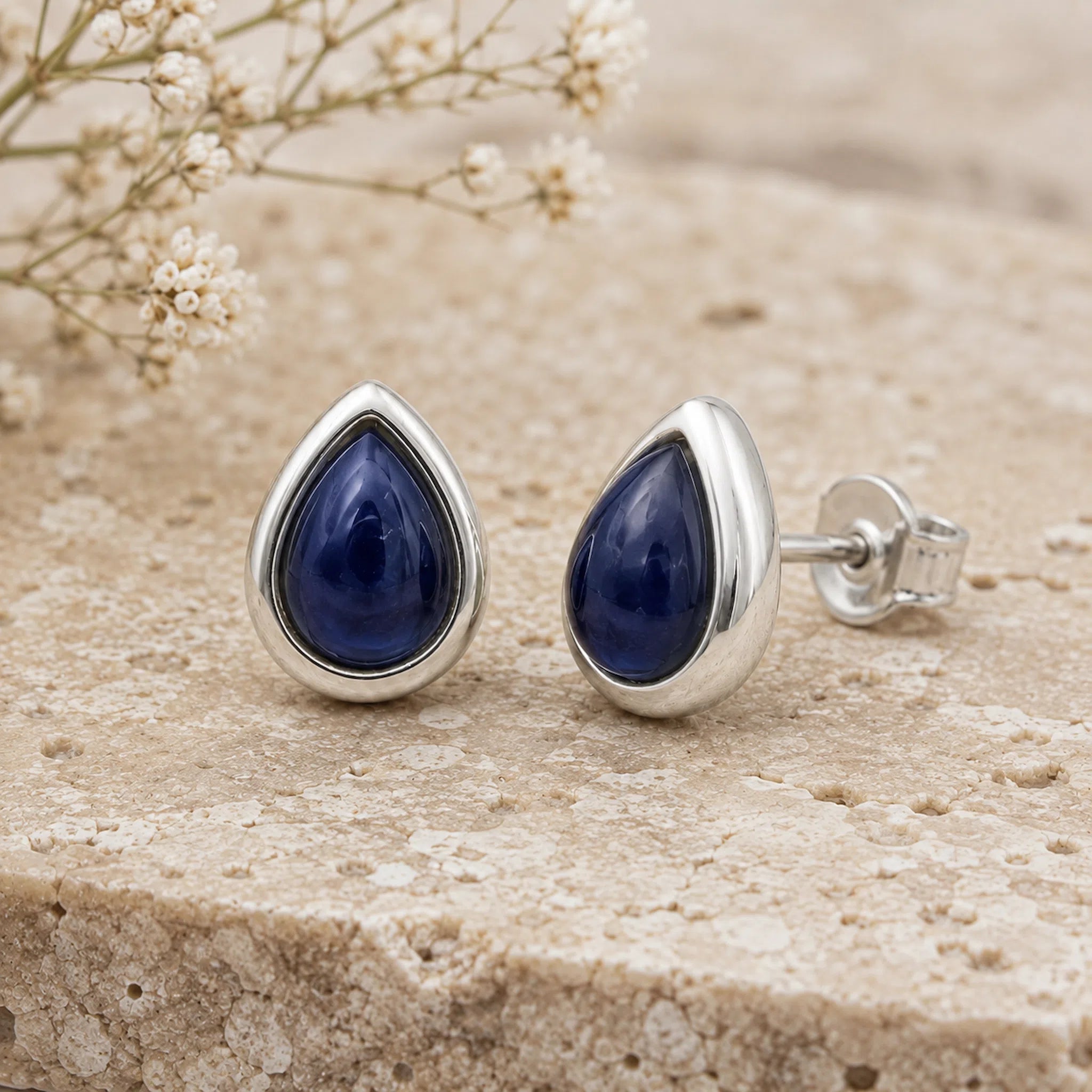 A pair of September Birthstone natural Sapphire Earrings - silver teardrop stud earrings with deep blue gemstone centers, displayed on a textured beige stone surface with small white flowers in the background.