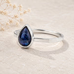 september birthstone natural sapphire ring - A silver ring featuring a teardrop-shaped dark blue gemstone with a cloudy, starry pattern, displayed on a light beige textured surface with dried flowers in the background.