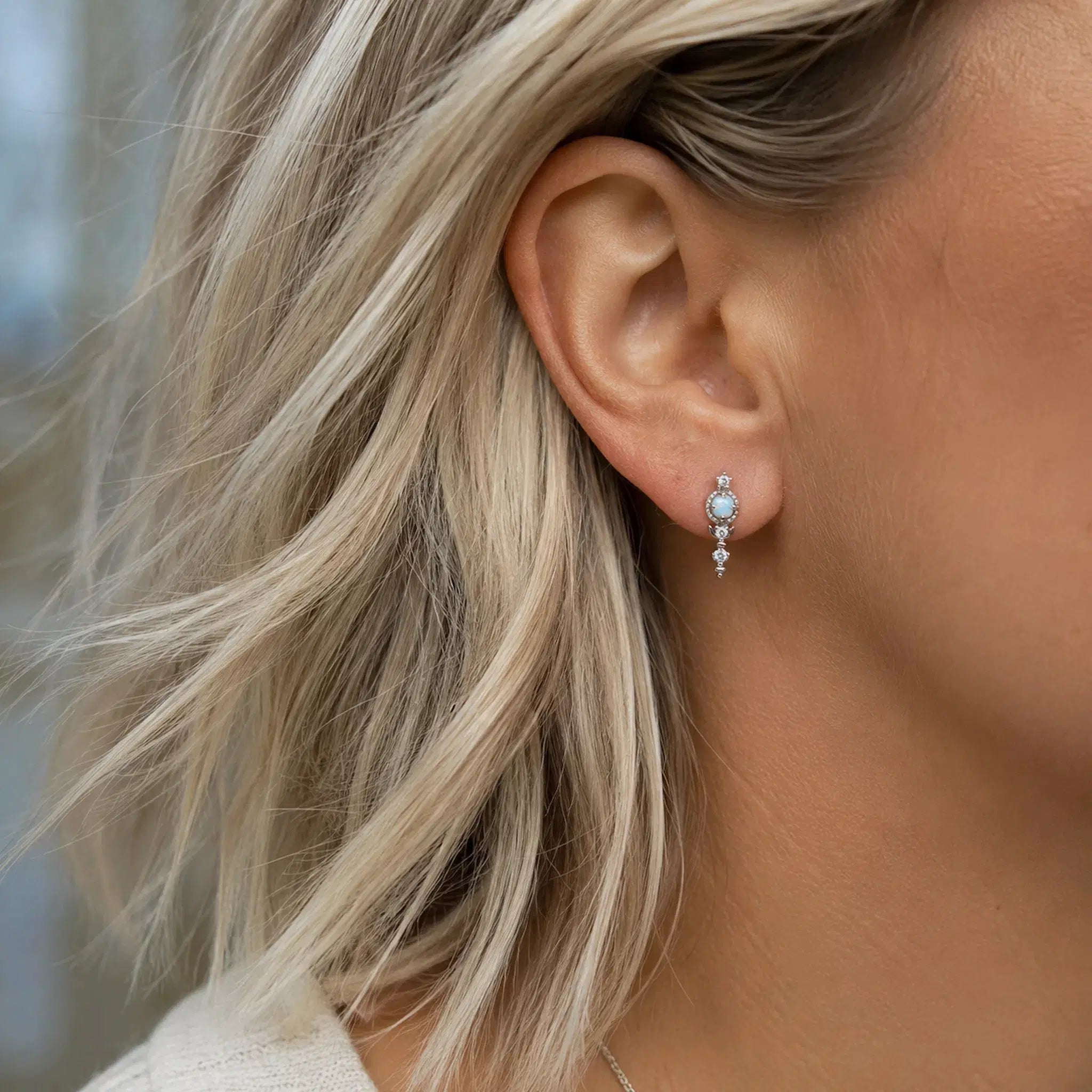 Close-up of an ear wearing a silver earring with a opal and CZ gemstones, blonde hair.