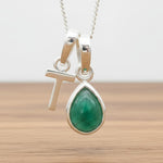 Silver Initial T necklace with a teardrop-shaped Emerald gemstone pendant hanging above a rustic wooden surface
