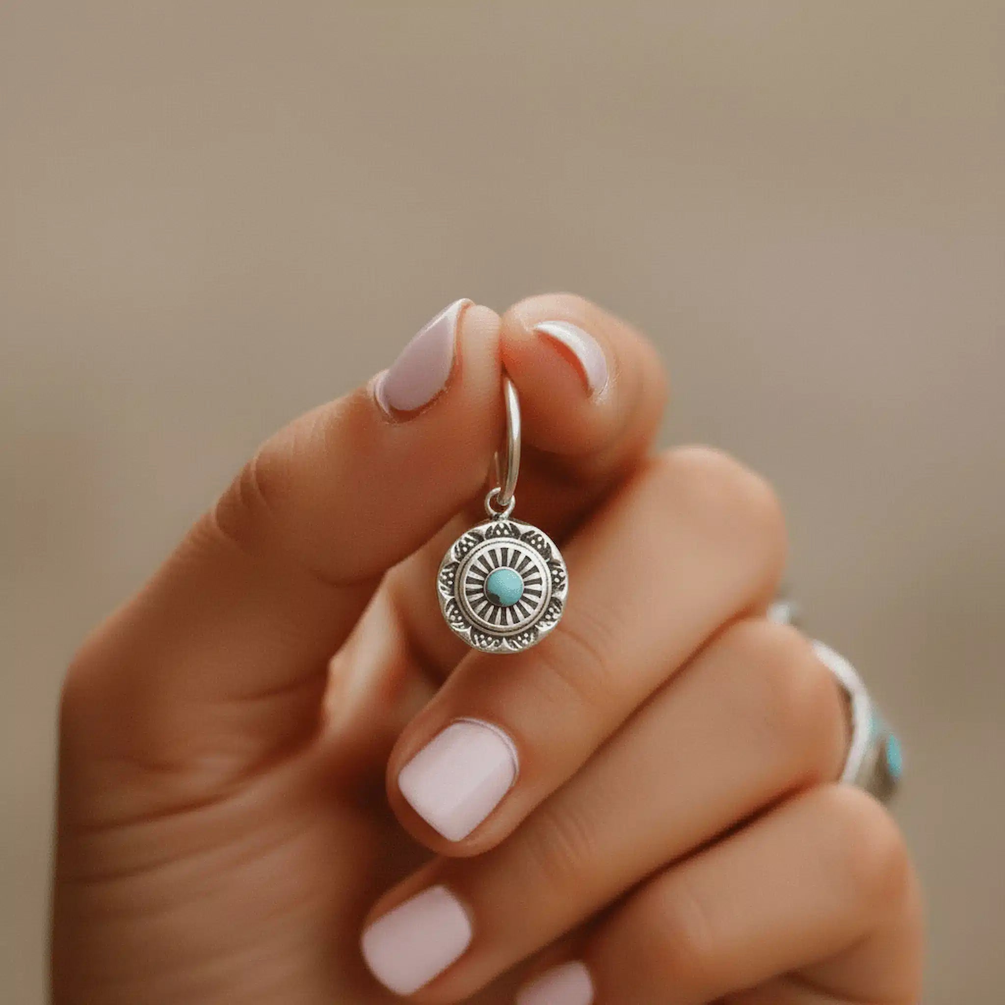 Hand holding a silver earring with a turquoise stone against a beige background