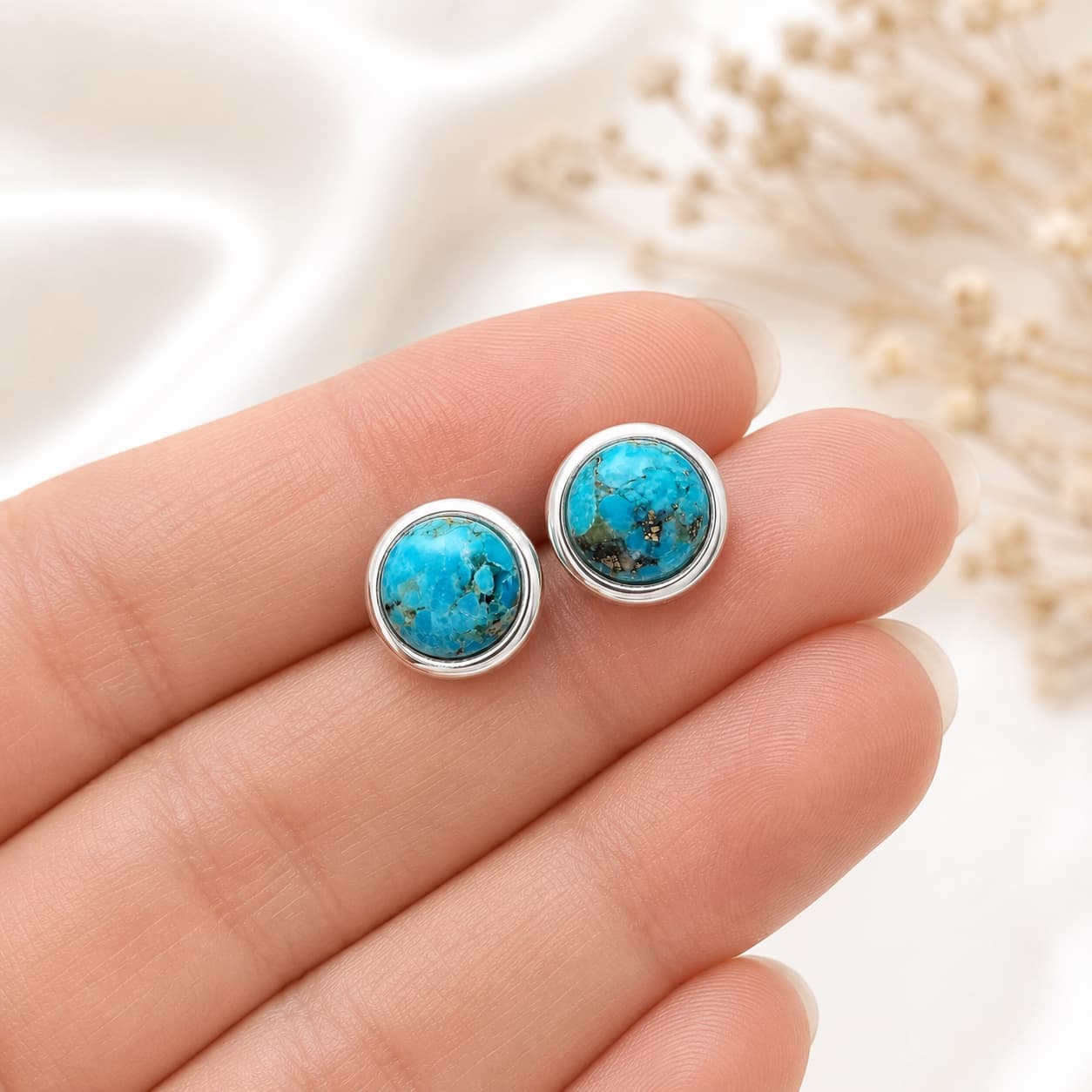 A pair of round turquoise stud earrings with silver bezel settings, held on a person's fingers against a soft white background with dried flowers.