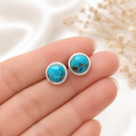 A pair of round turquoise stud earrings with silver bezel settings, held on a person's fingers against a soft white background with dried flowers.