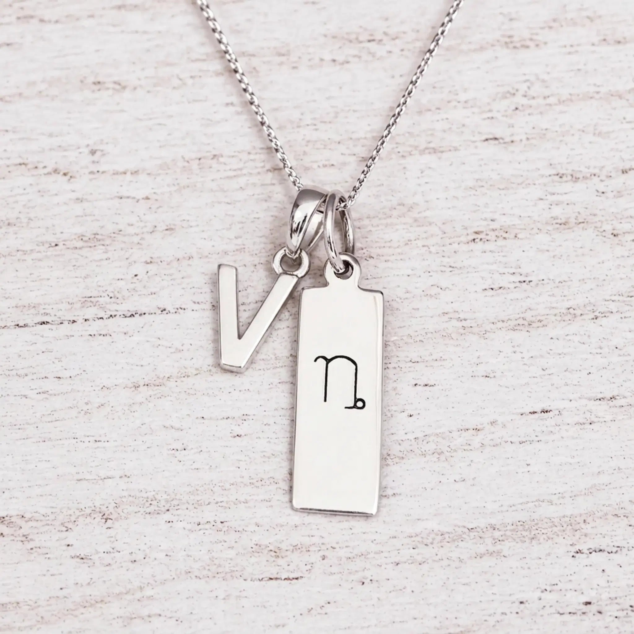 sterling silver initial V pendant and capricorn necklace on a fine silver chain against a light wooden background