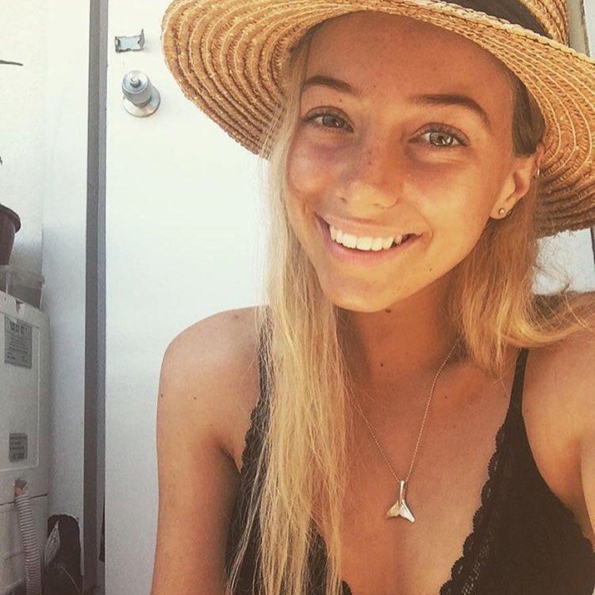 Woman wearing a straw hat and black top with a silver whale tail necklace, smiling outdoors.