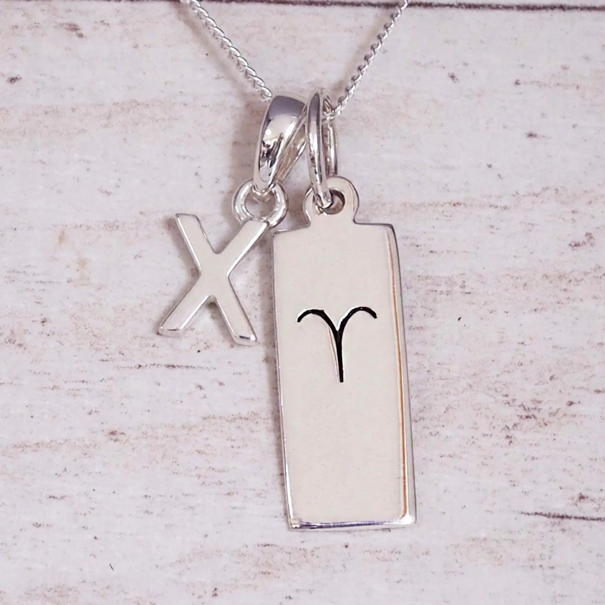 Sterling Silver X Initial pendant and Aries necklace on a fine silver chain on a light wooden background