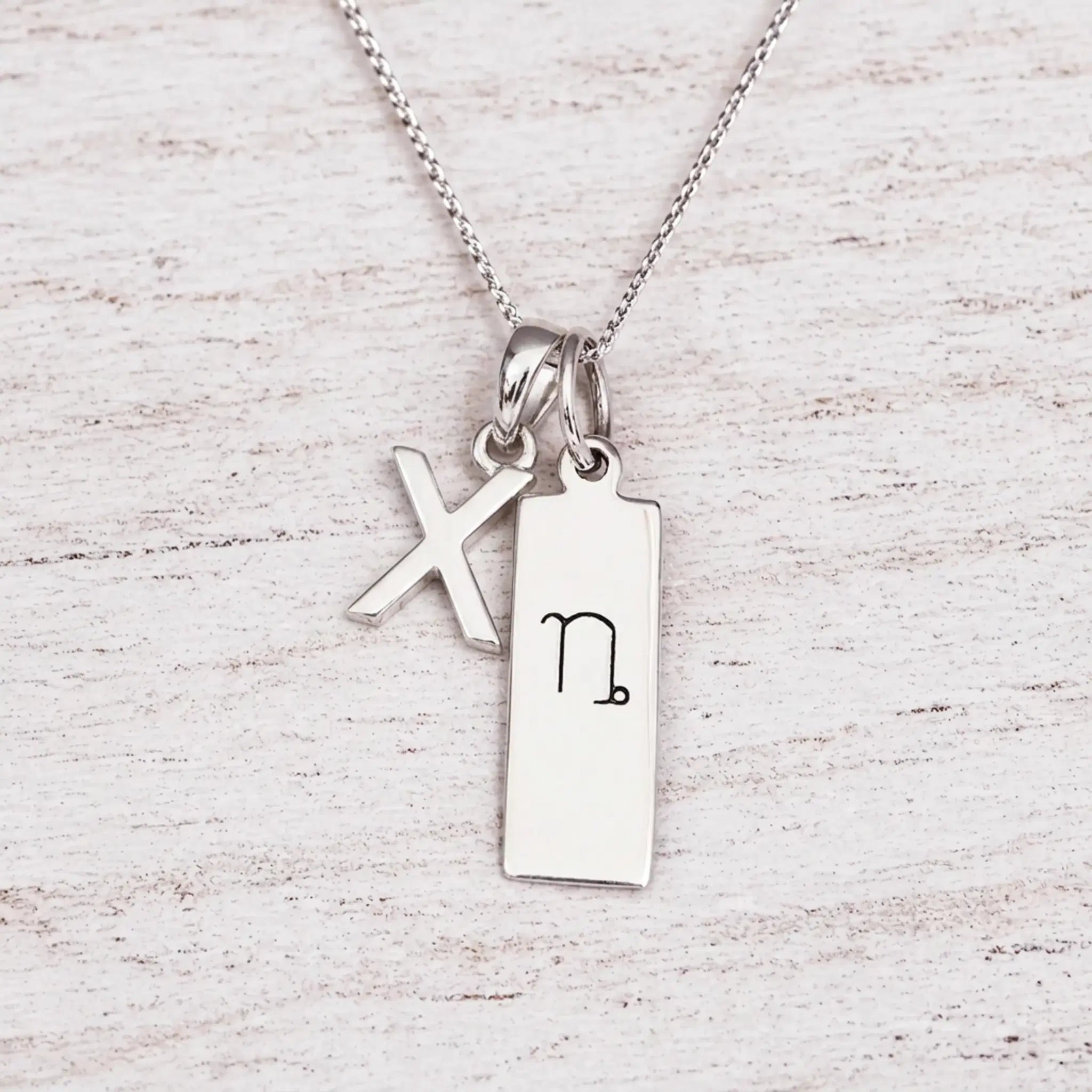 sterling silver initial X pendant and capricorn necklace on a fine silver chain against a light wooden background