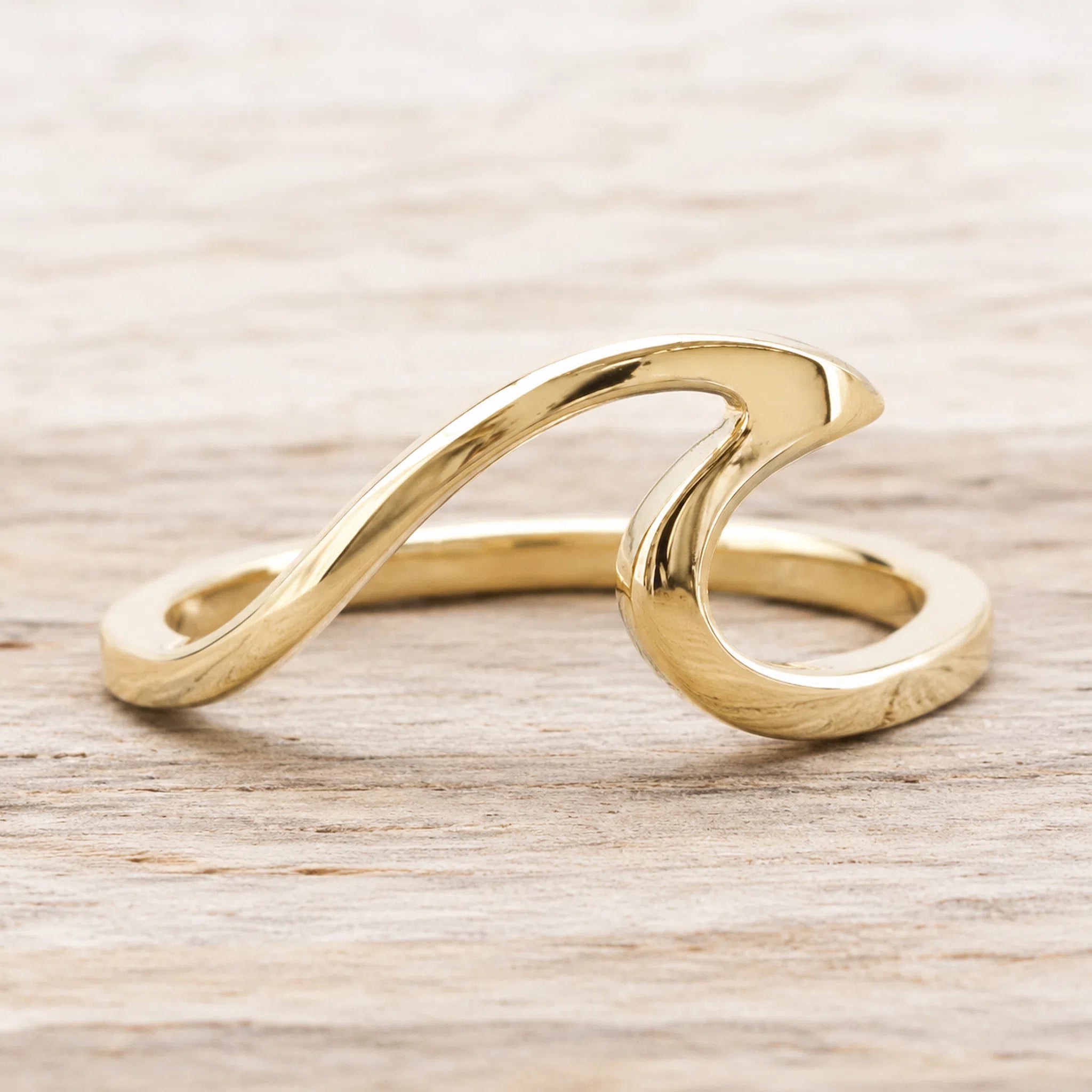 A polished gold wave-shaped ring resting on a light-colored wooden surface.