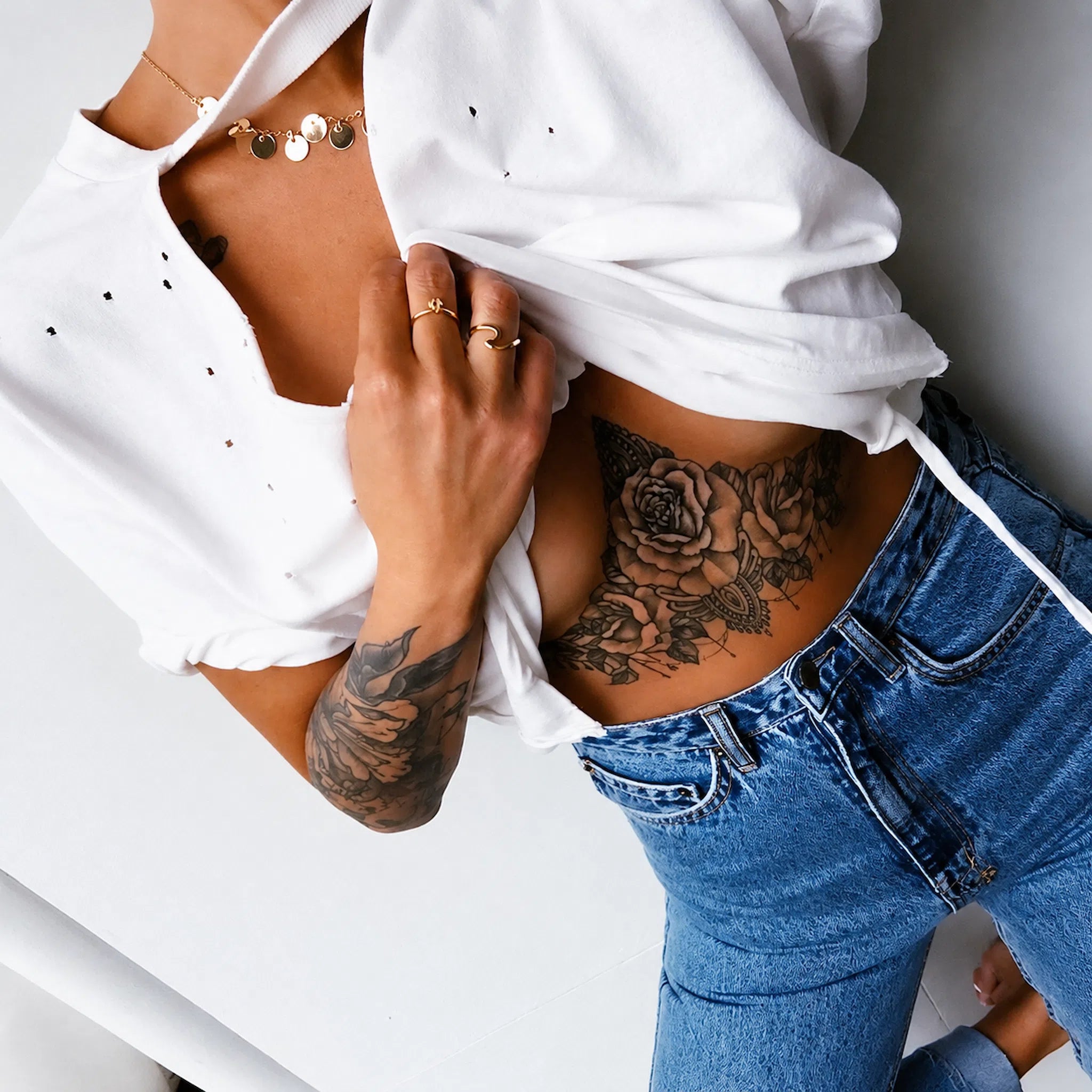 A close-up lifestyle shot of a person wearing a white short-sleeve top with small black dot details and blue jeans, lifting the shirt to show a floral rose tattoo on their lower back and arm. They are wearing gold rings and a gold coin necklace.