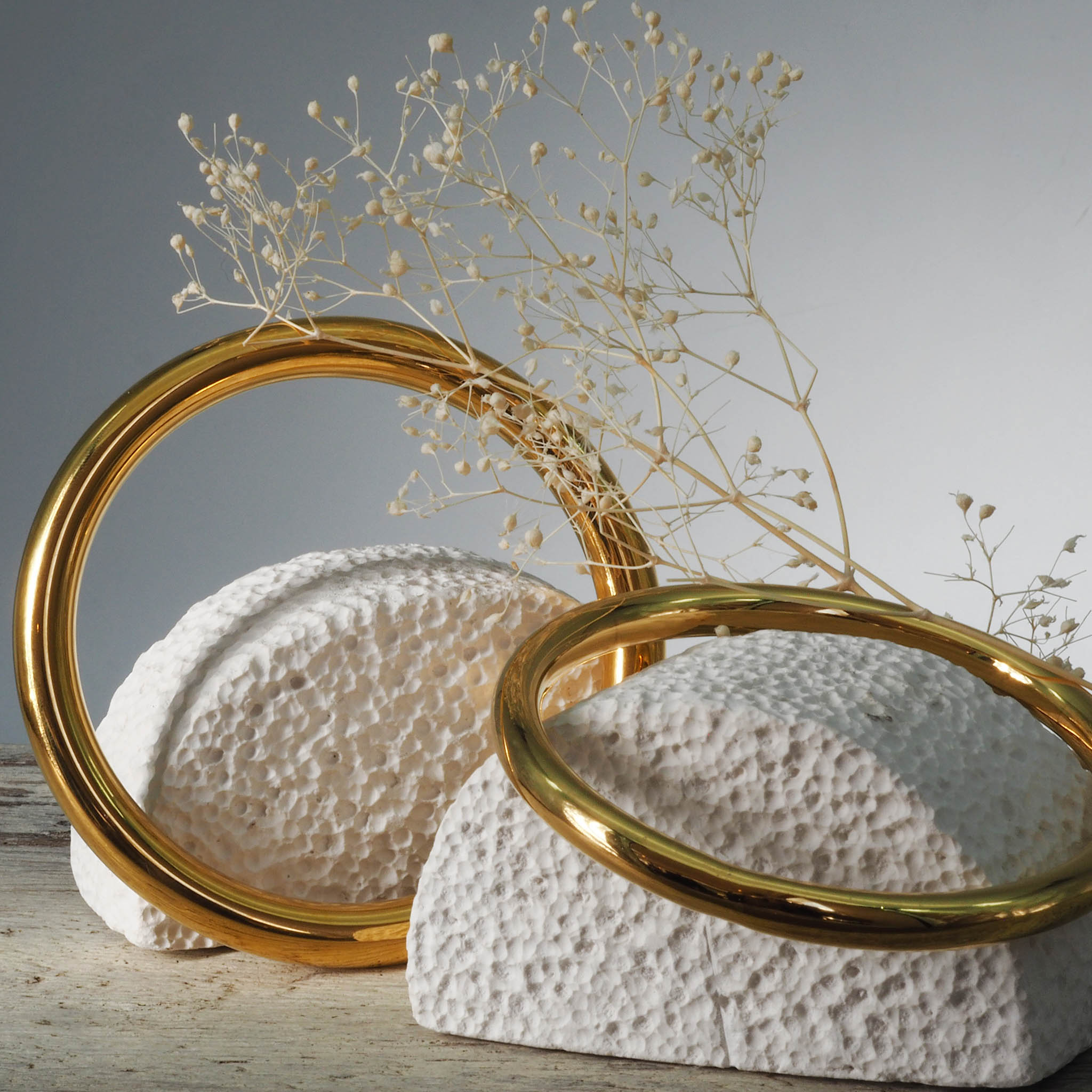 Decorative items with two gold stainless steel bangle bracelets in polished finish on a textured white bases on a wooden surface.